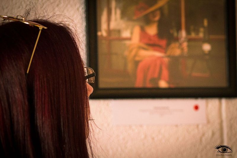 a female figure with long brown hair looks on a picture of a woman in a large hat and a red dress that is framed on the wall